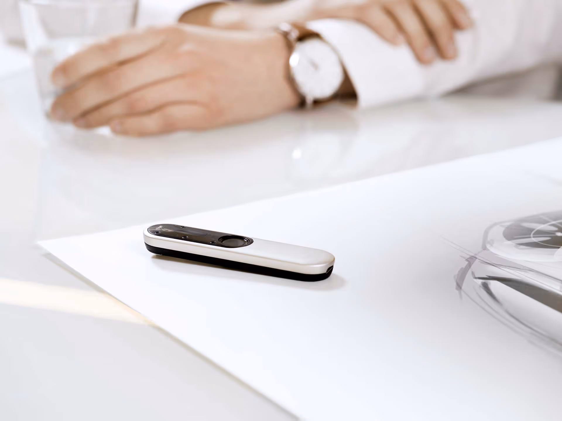A person sits at a table holding a glass, with a small device resting on some papers.