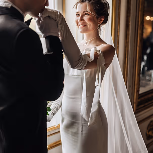Bride in a white wedding dress smiling as groom in a black suit kisses her gloved hand indoors.