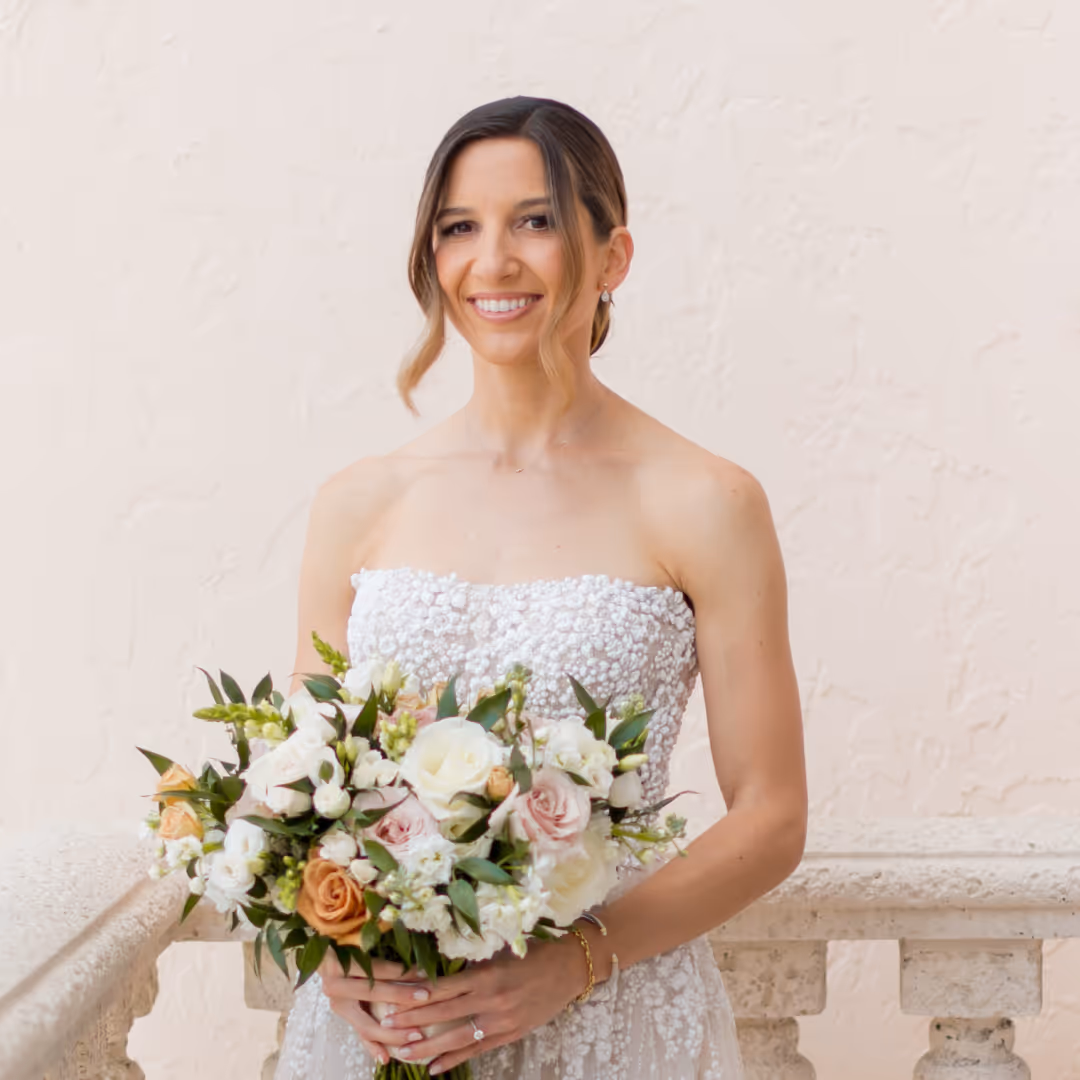 Smiling bride in a strapless white floral wedding dress holding a bouquet of white and peach flowers with greenery.