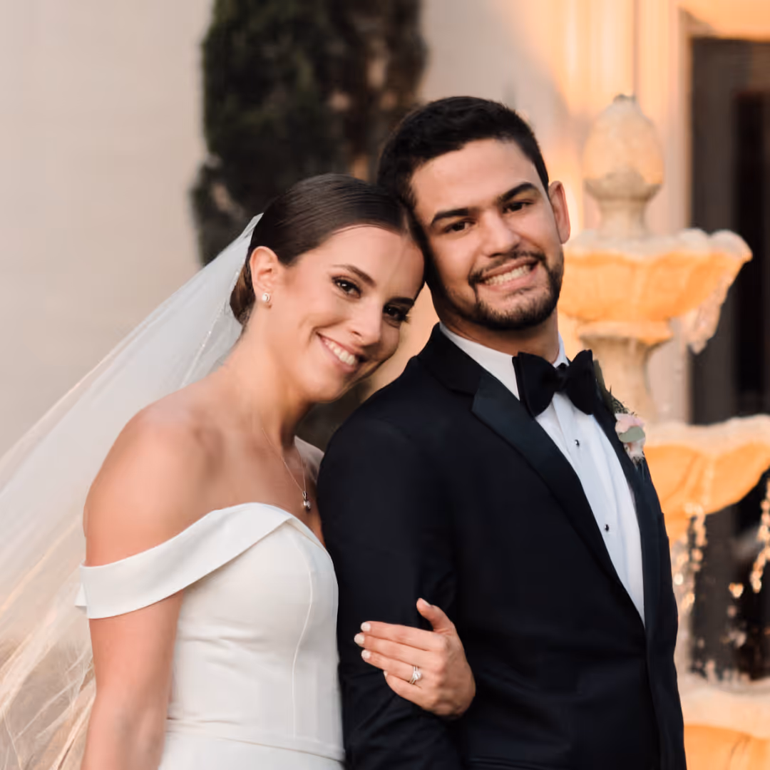Bride in an off-shoulder white wedding dress and veil smiles leaning on groom in black tuxedo in front of a stone fountain.