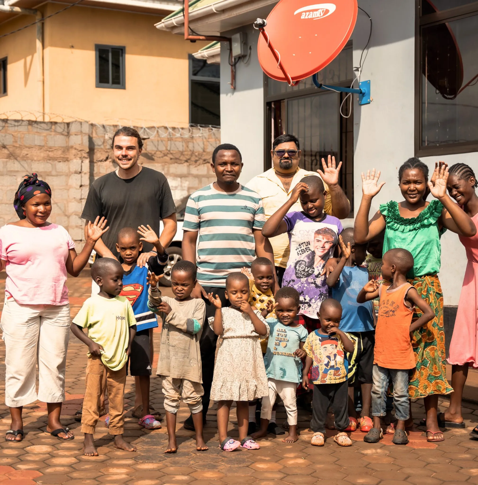 En gruppe smilende voksne og barn står utendørs foran en bygning med en rød parabolantenne.