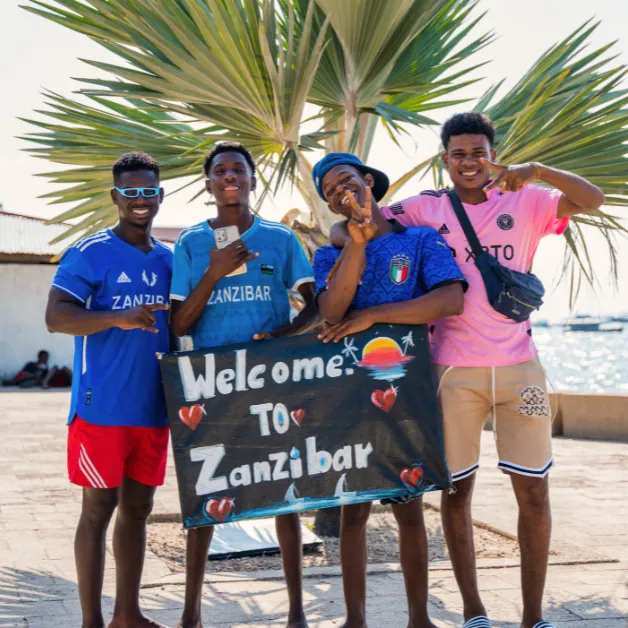 Fire unge menn står sammen utendørs, holder et skilt med teksten «Velkommen til Zanzibar» og lager et fredstegn.