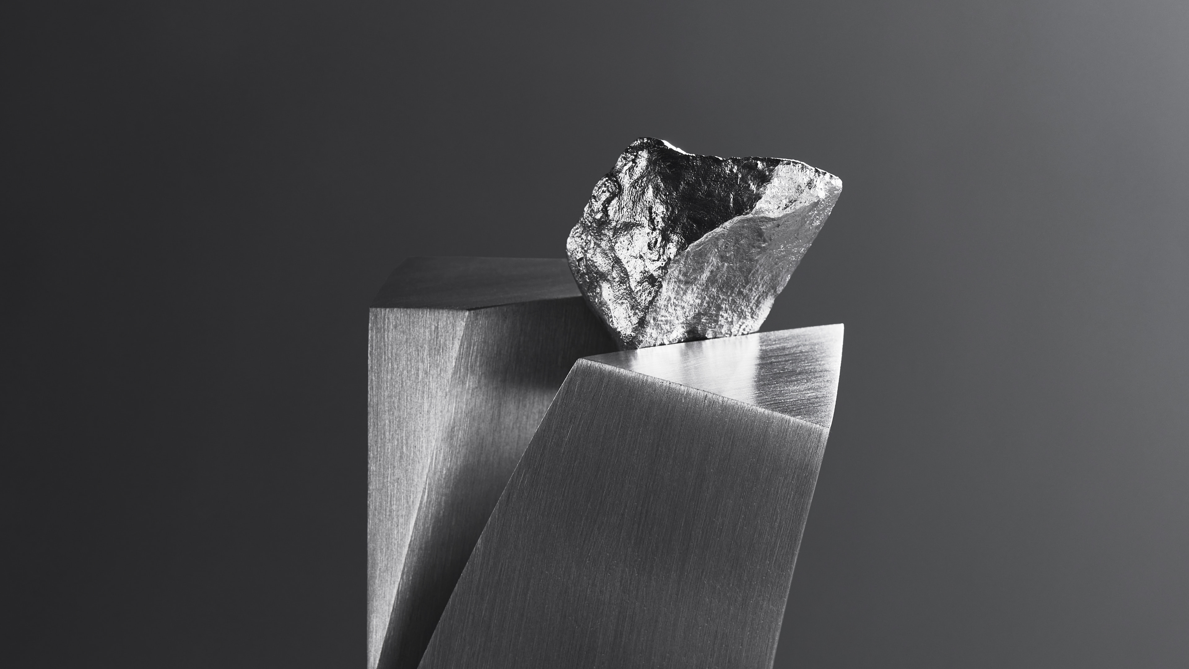 Wide crop of the rough metal rock resting on the angled faces of the brushed metal slabs, on a dark grey background.