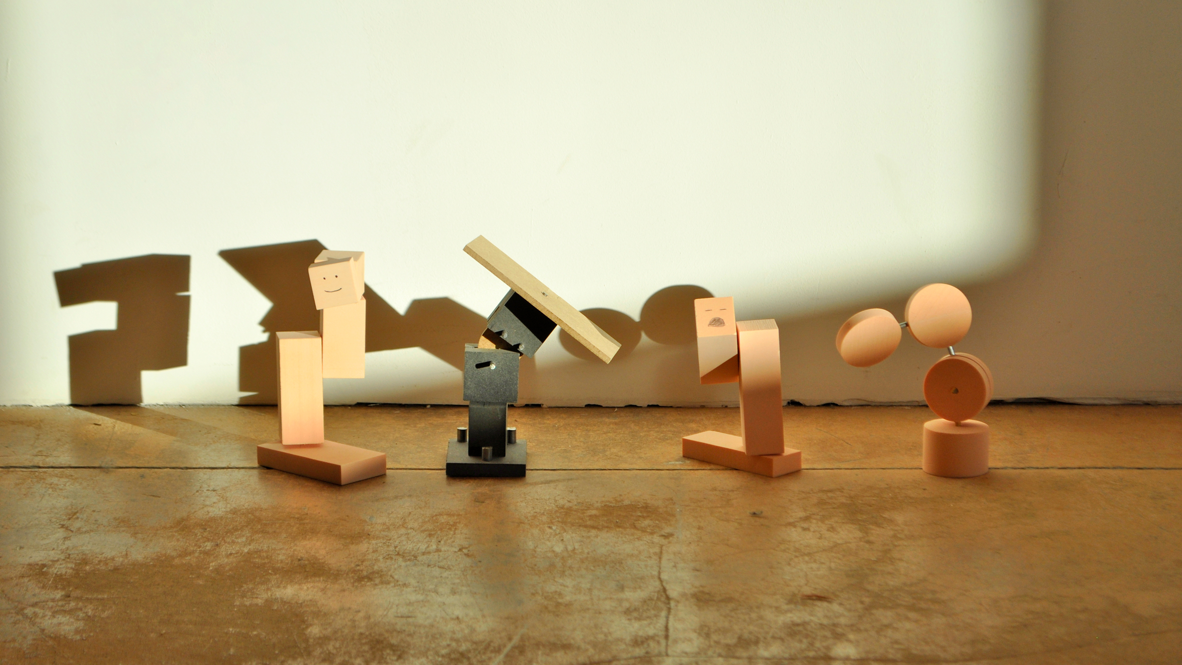Early prototypes: four small foam, wood and metal figures with simple faces, one smiling and one with a moustache, posed on a concrete floor and casting long shadows on a white wall.
