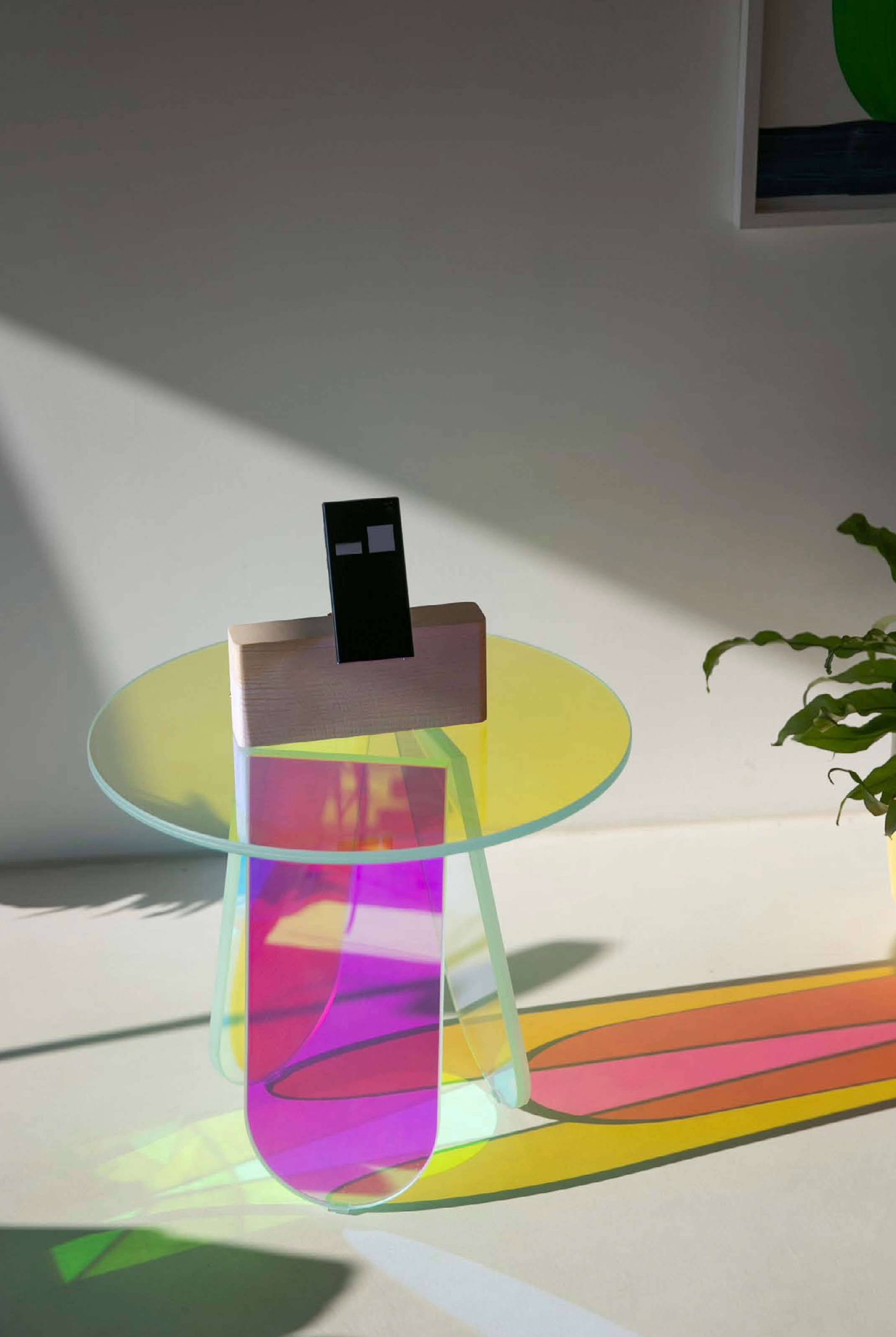 Phone with two white rectangular eyes in its pale wooden base on a round, iridescent acrylic side table, with pink, yellow and green tints and coloured light on the floor.