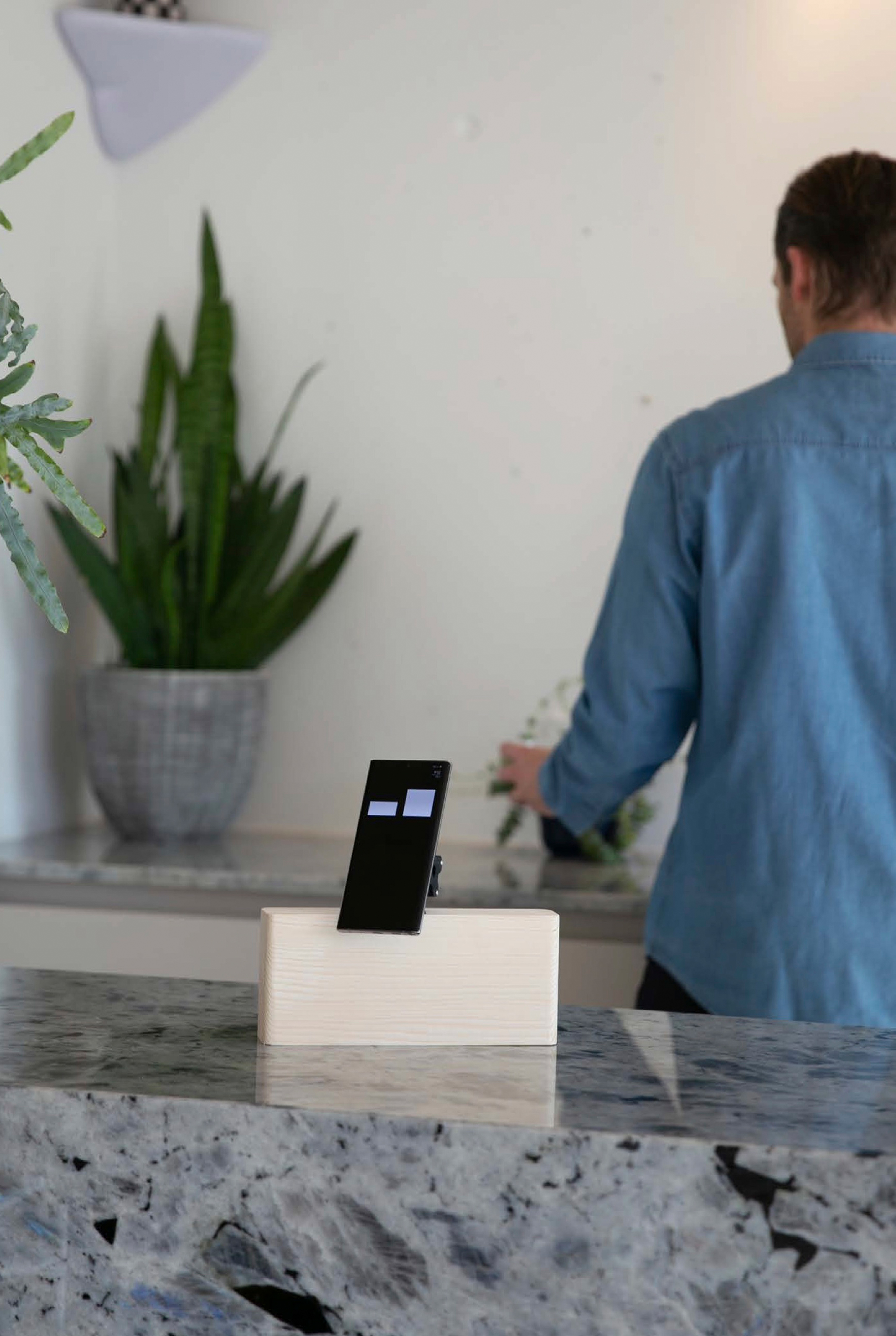 Phone assistant with two white rectangular eyes standing in its pale wooden base on a granite counter, with a man in a blue shirt and a potted plant behind.