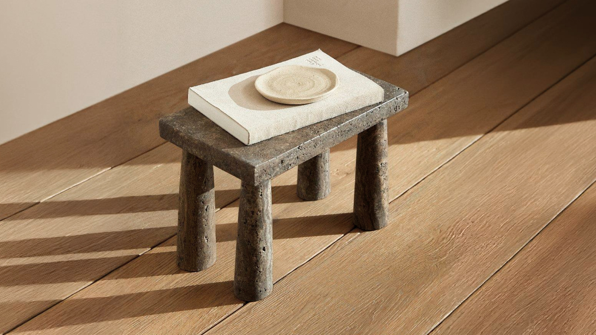Tap stool in dark travertine on an oak floor in raking sunlight, with a cream book and a small ceramic dish on top.