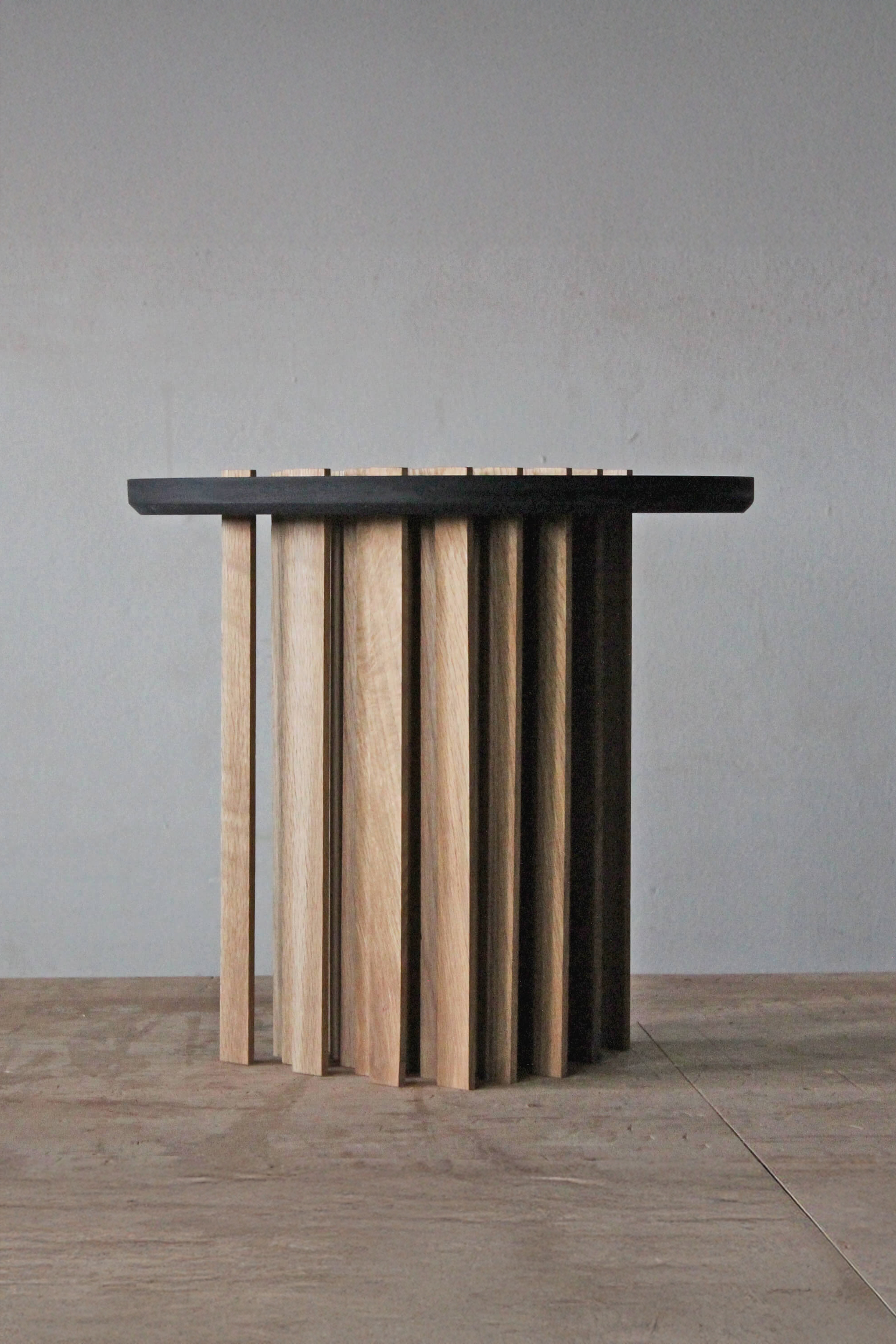 Small Bohr table with a round black top on a cluster of upright oak slats of different widths, against a grey wall.