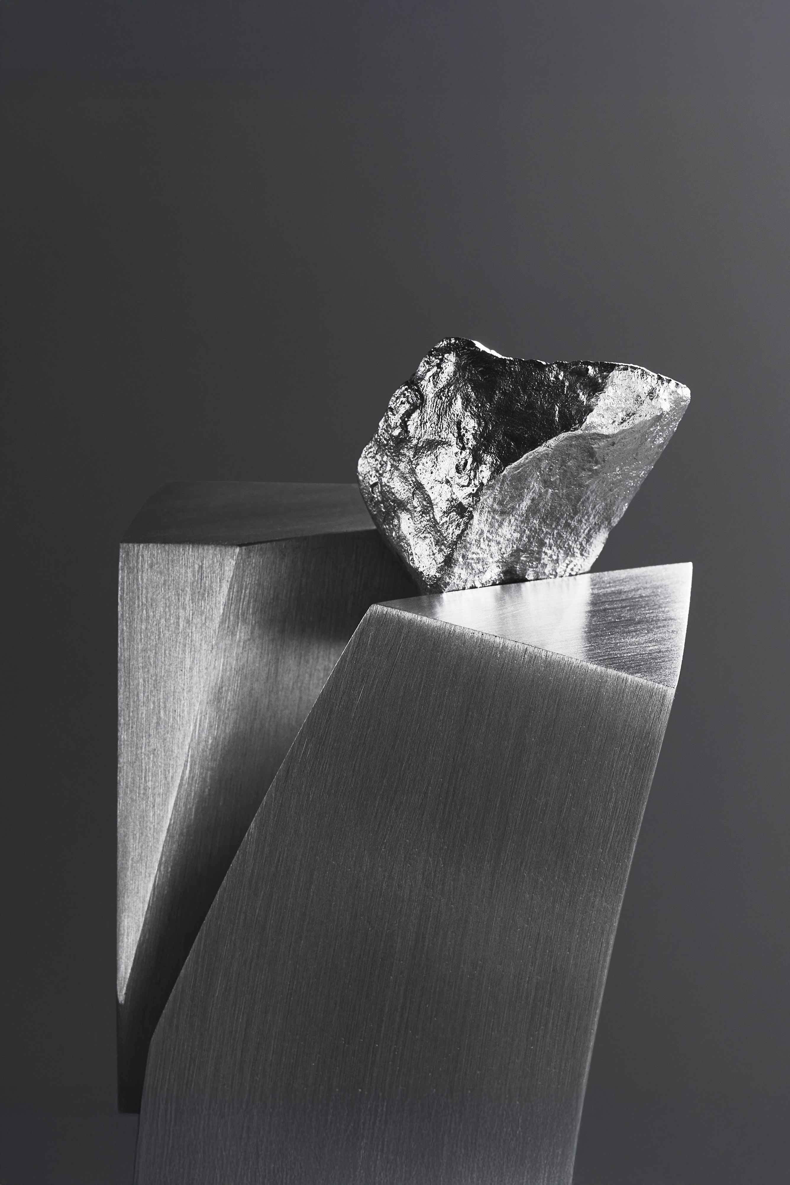 Eternal Glance sculpture in close-up: a rough, silvery rock balanced on the angled top of two brushed metal slabs against a dark grey background.