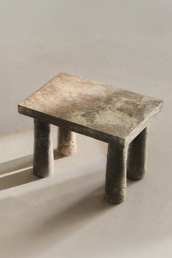 Tap stool from above, with a rectangular travertine top on four tapered round legs, and sunlight and shadow across a pale floor.