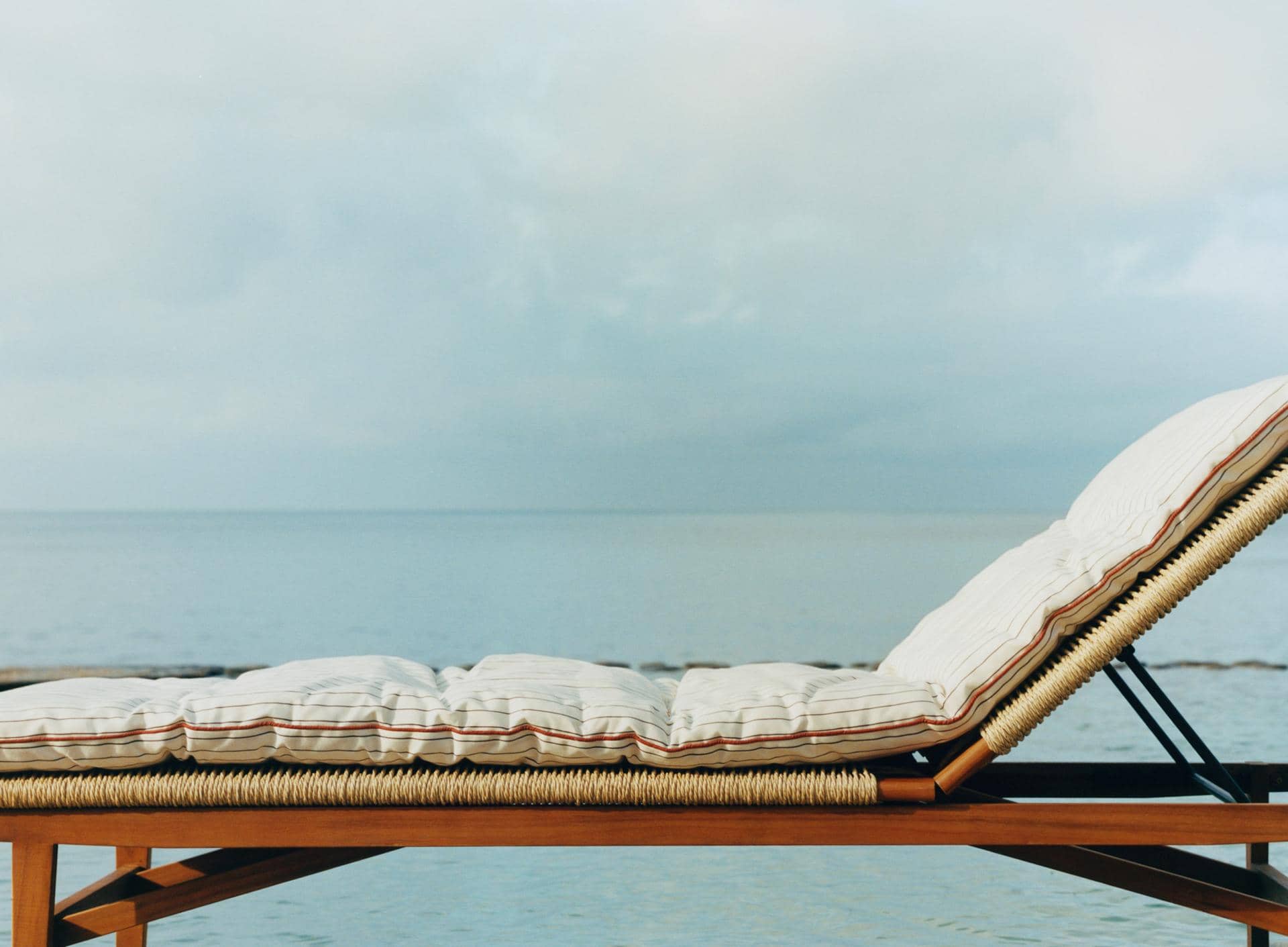 Cord lounger with a cream cushion edged in red stripes, back raised, facing a calm sea and cloudy sky.