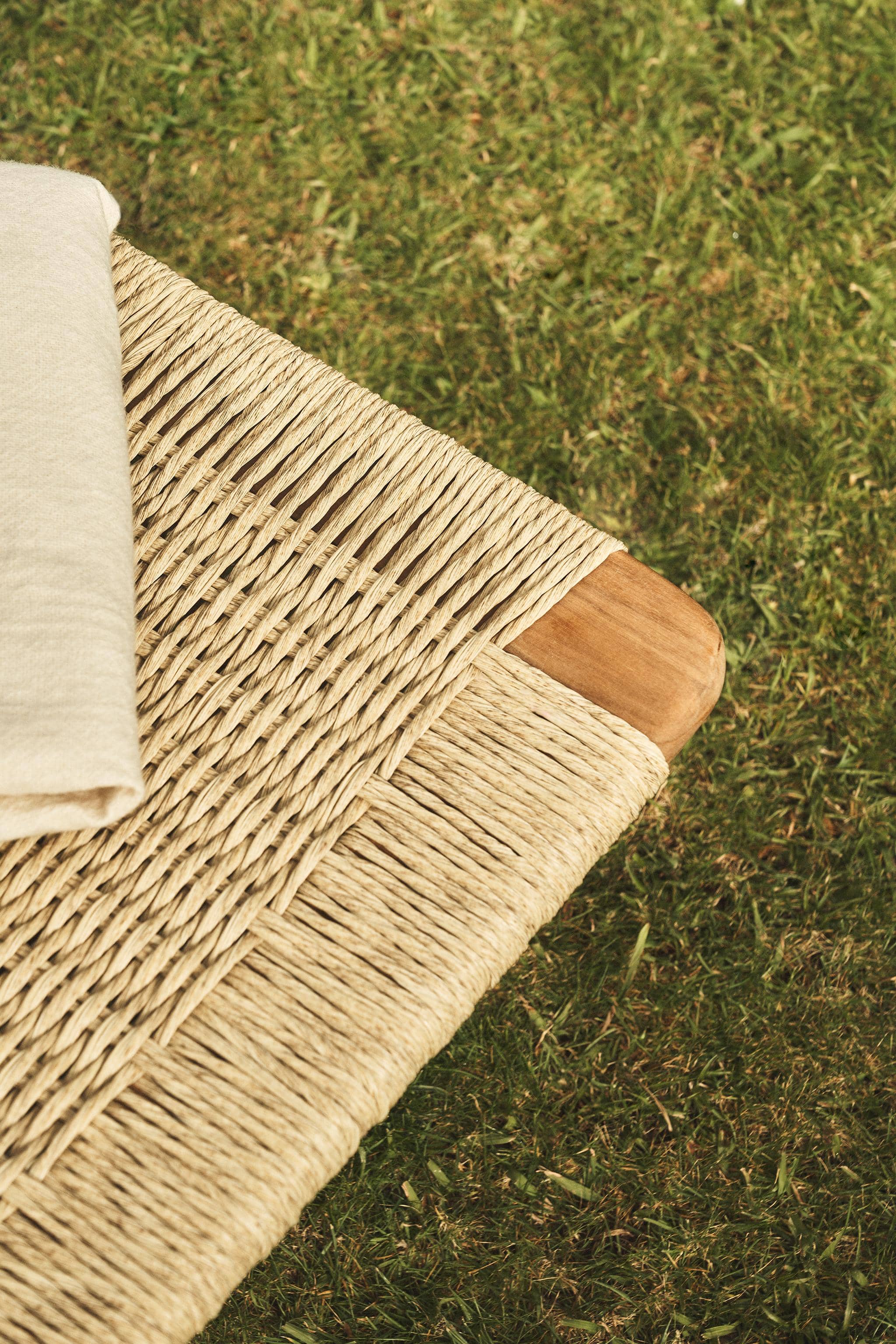 Detail of a woven cord seat meeting a rounded teak corner, with a folded cream cloth and grass beneath.