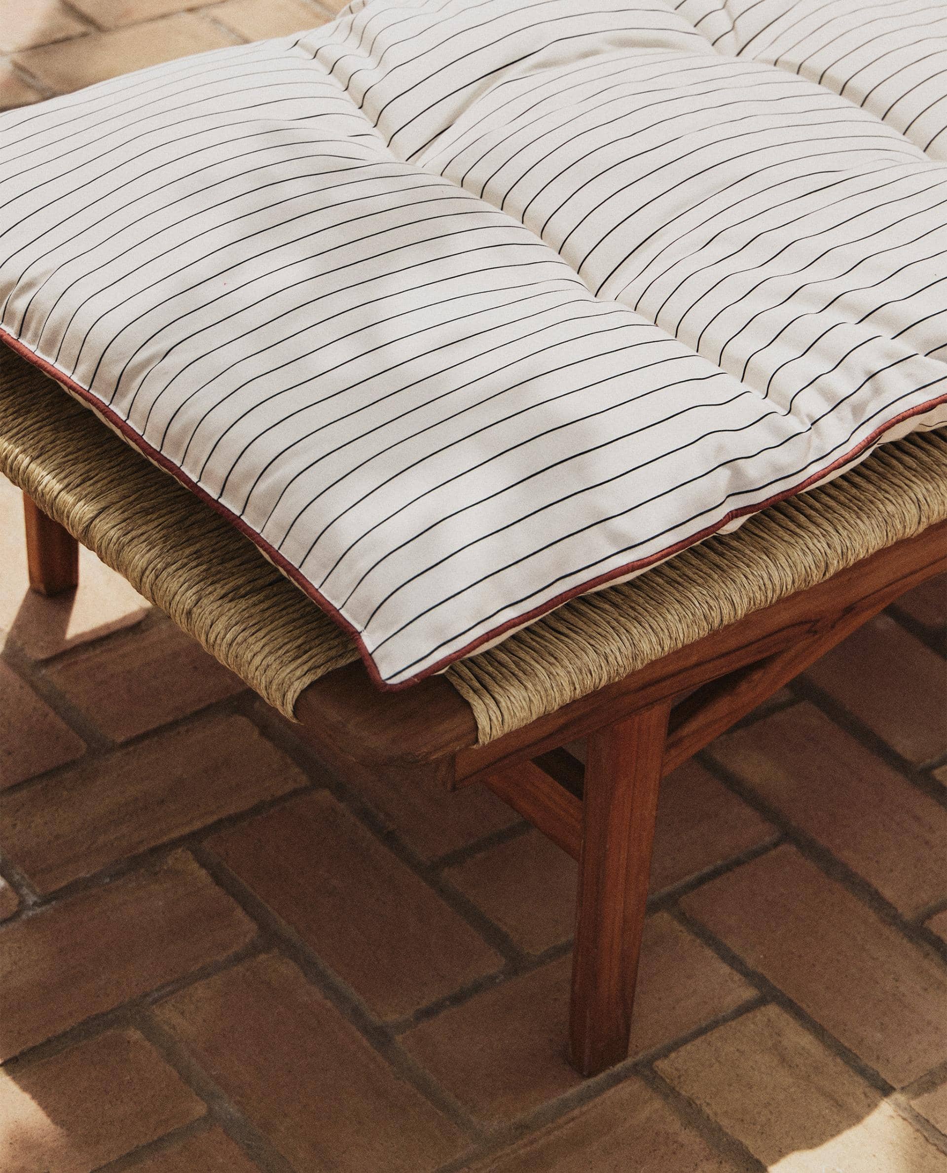 Close-up of a cream cushion with thin dark stripes and red piping over the woven cord seat of the teak lounger, on a terracotta brick floor.