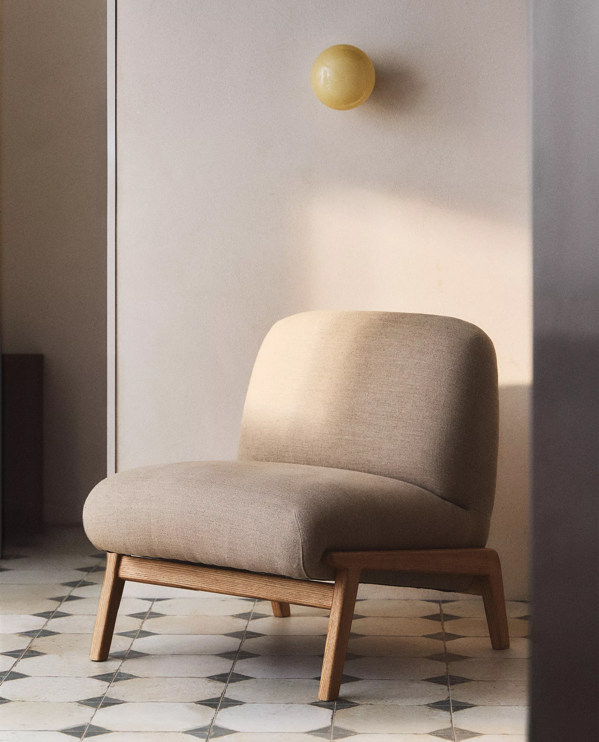 Beige linen Camilla chair with a light oak frame in a sunlit room, on a black and white diamond-tiled floor, with a round yellow glass wall light behind it.