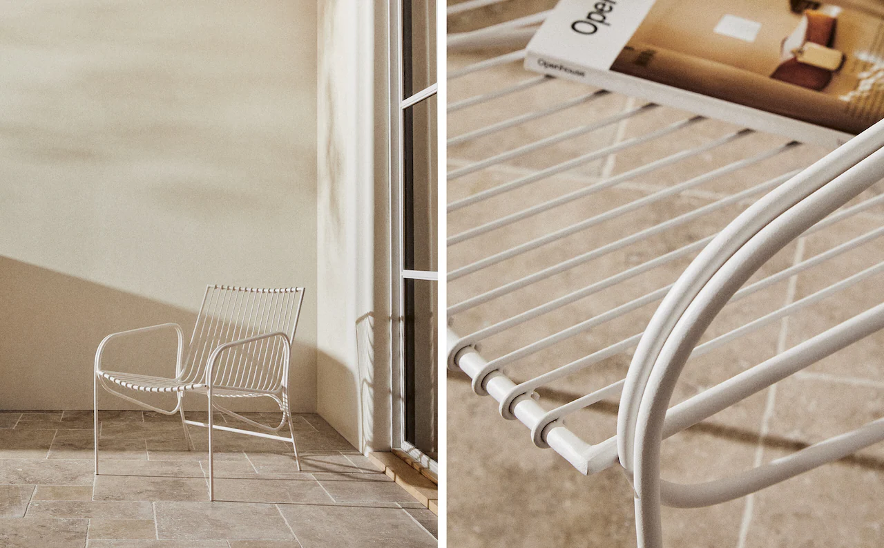 White Cala armchair in a corner beside a glass door, on pale stone tiles, with slender rods for the back and seat and rounded armrests.