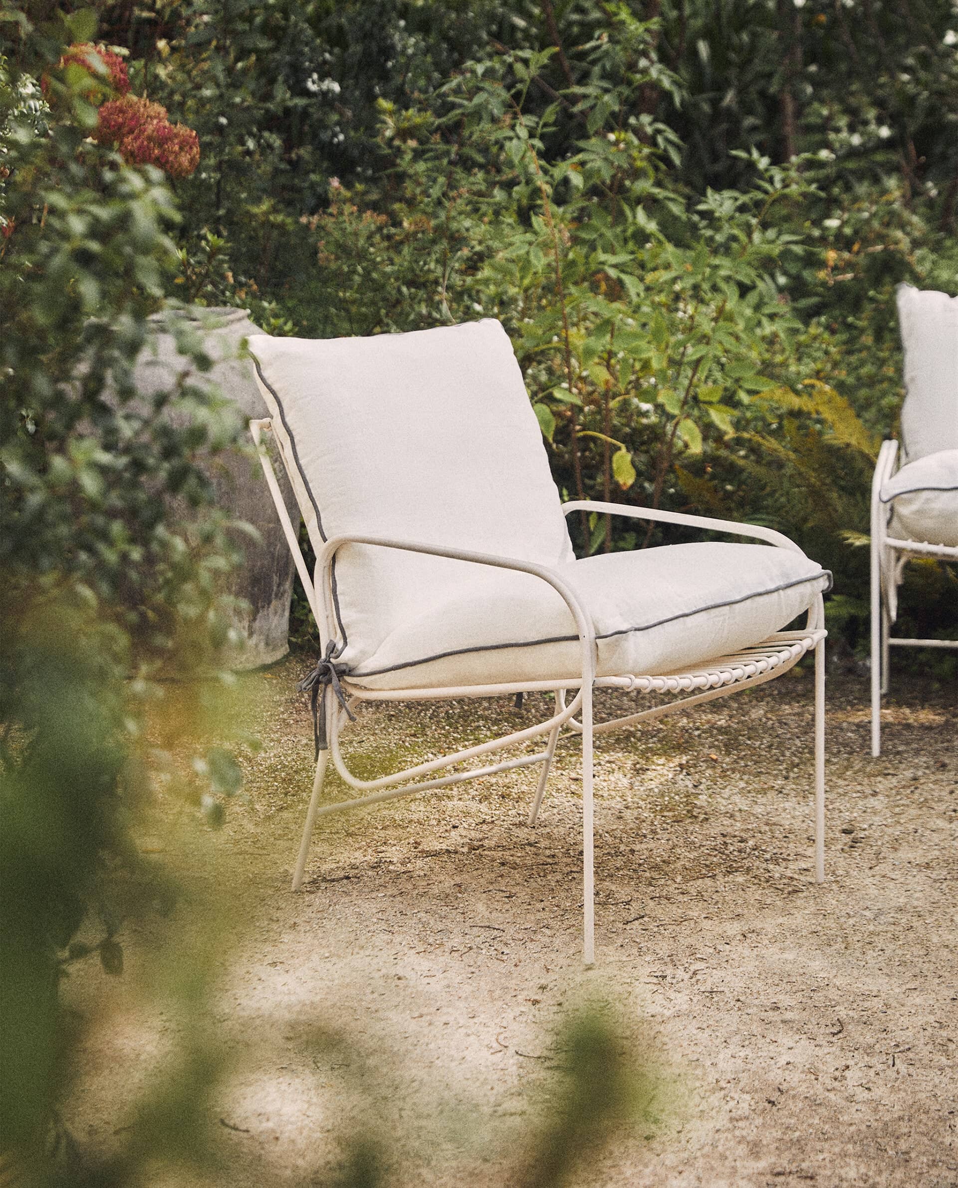 White Cala armchair with thick cream cushions piped in grey and tied at the back, on gravel in a leafy garden.