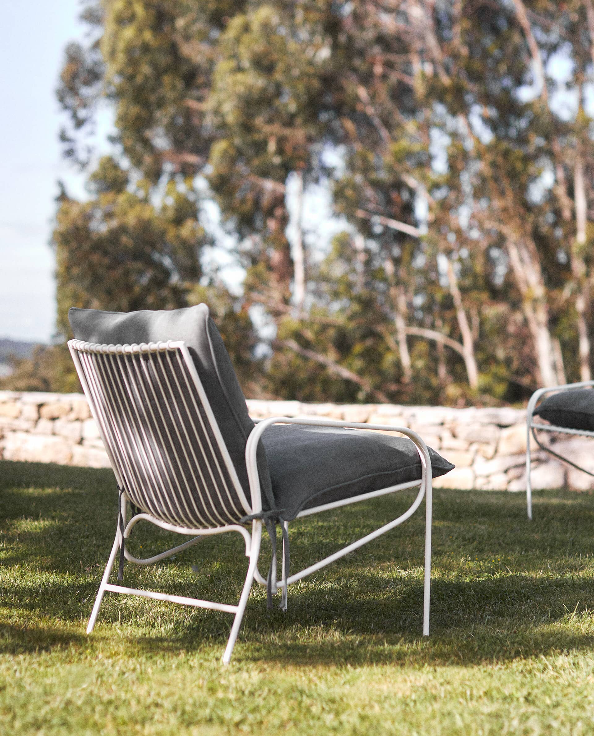 Rear view of a white Cala chair with a grey cushion on grass, in front of a dry stone wall and eucalyptus trees.