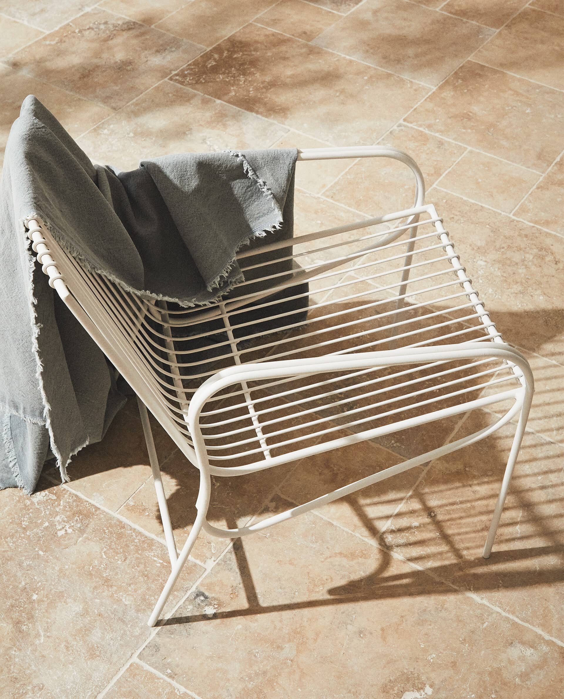 White Cala chair seen from above on honey-coloured stone tiles, with a grey fringed blanket over the back and a long shadow.