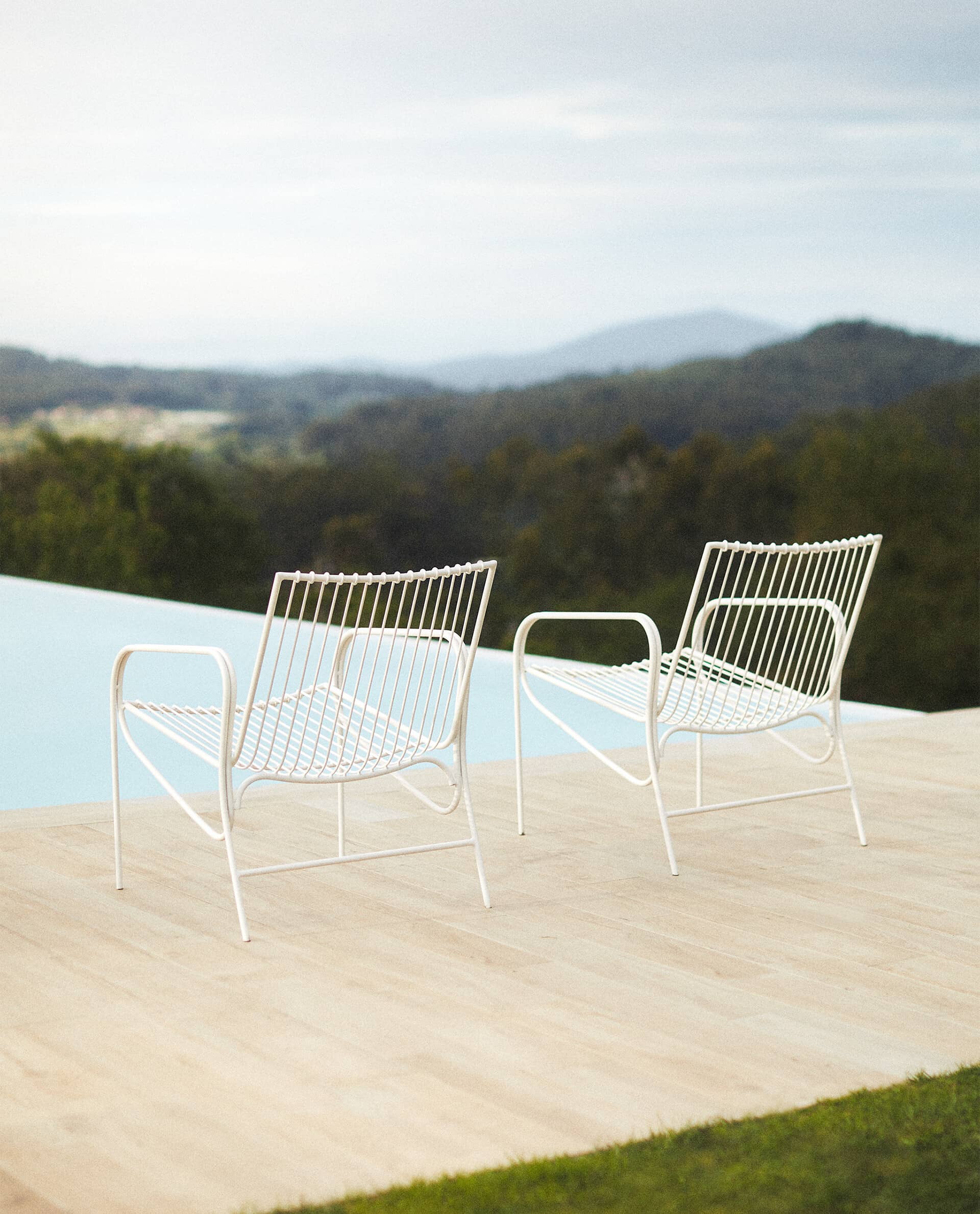 Two white Cala chairs side by side near the edge of a pool, with wooded hills behind.