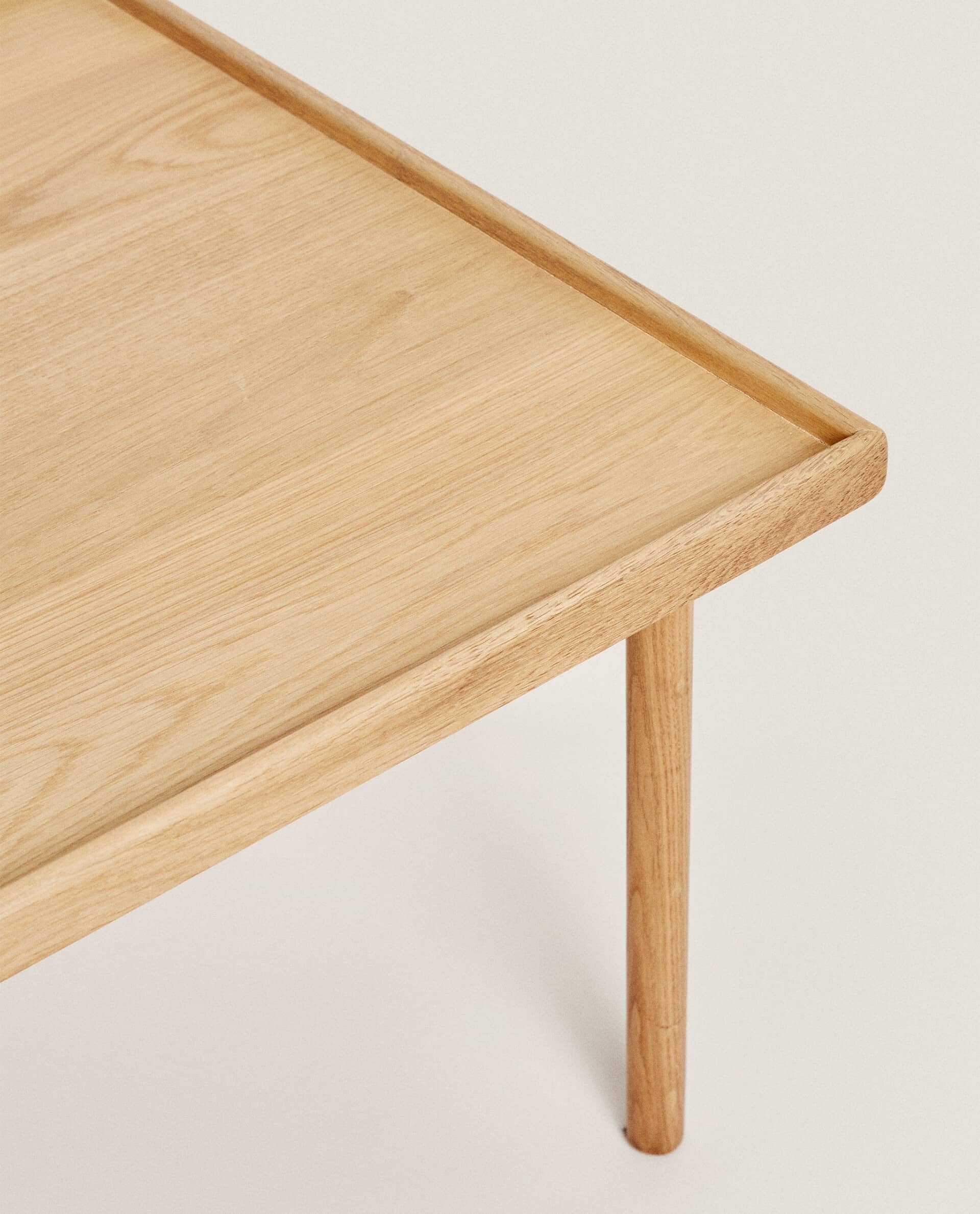 Corner detail of the Oat table, showing the oak grain, the raised rim and a slim leg meeting the underside of the top.