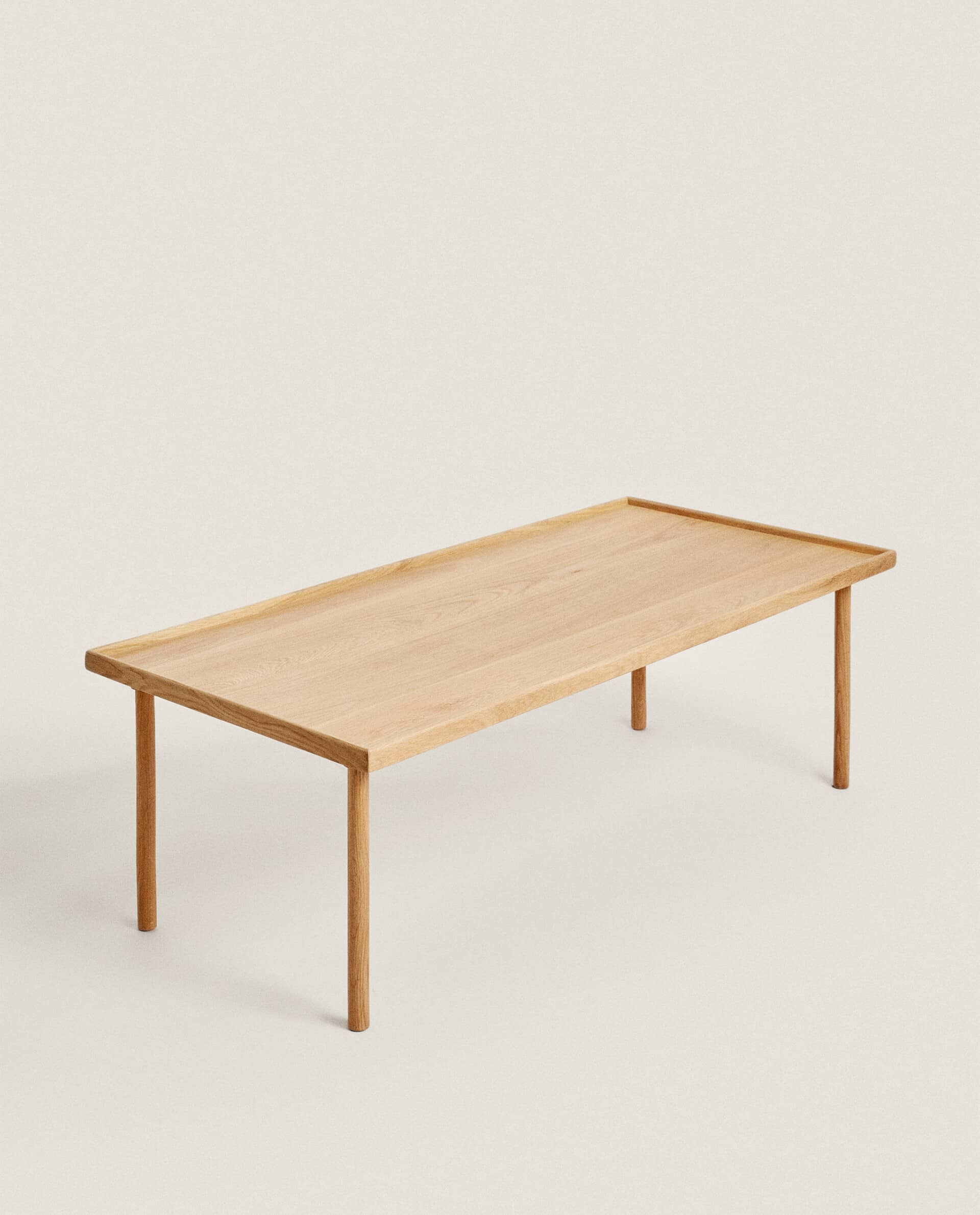 Three-quarter view of a long, low Oat table in light oak, with a raised rim and four slim round legs, on a plain pale background.
