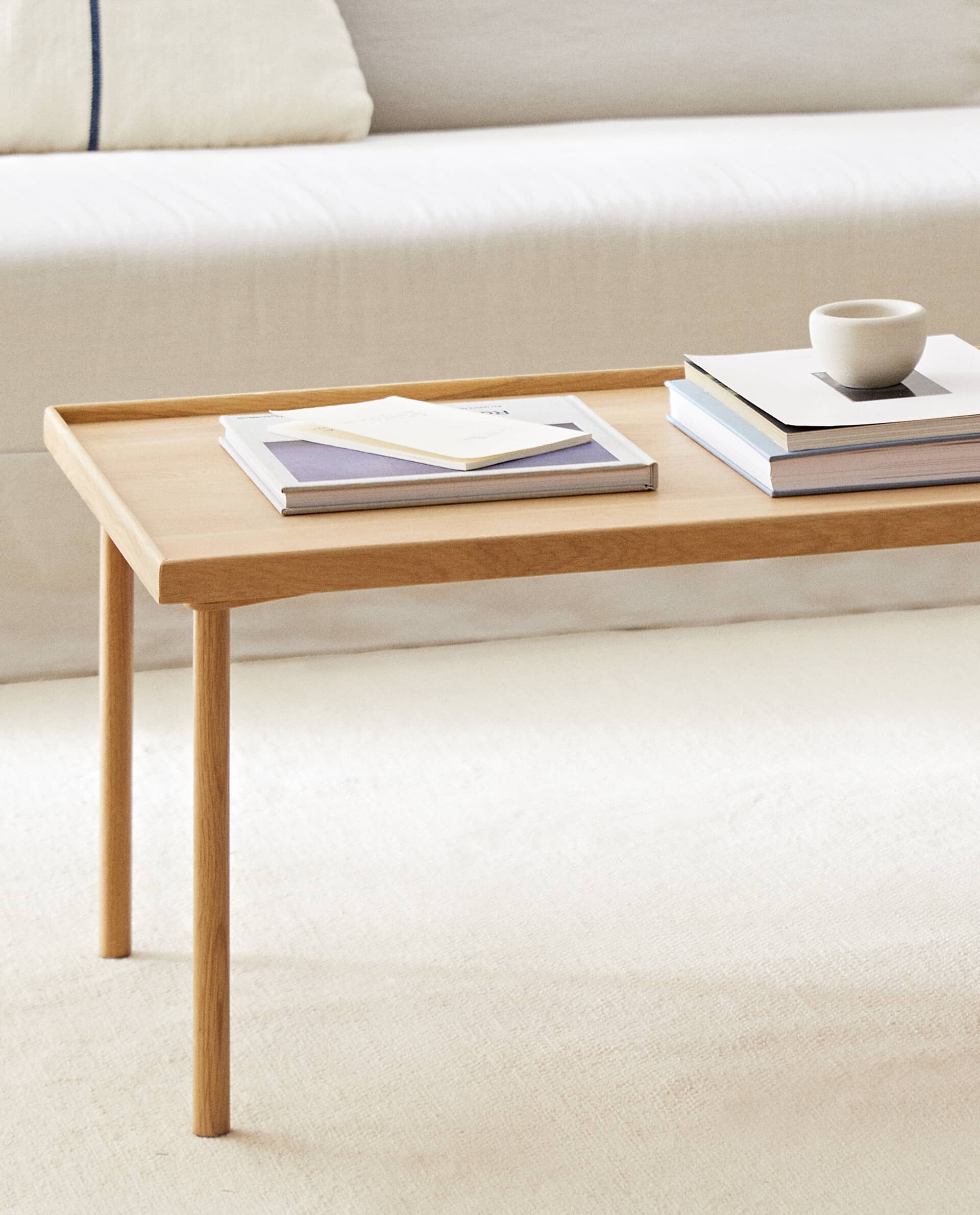 Oat rectangular table in light oak with a raised edge, holding books, a notebook and a small ceramic bowl, on a cream rug in front of a sofa.