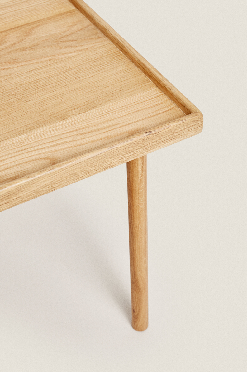 Close-up of an Oat table's corner in light oak, with a thin raised lip around the top edge and a slim round leg, on a cream background.