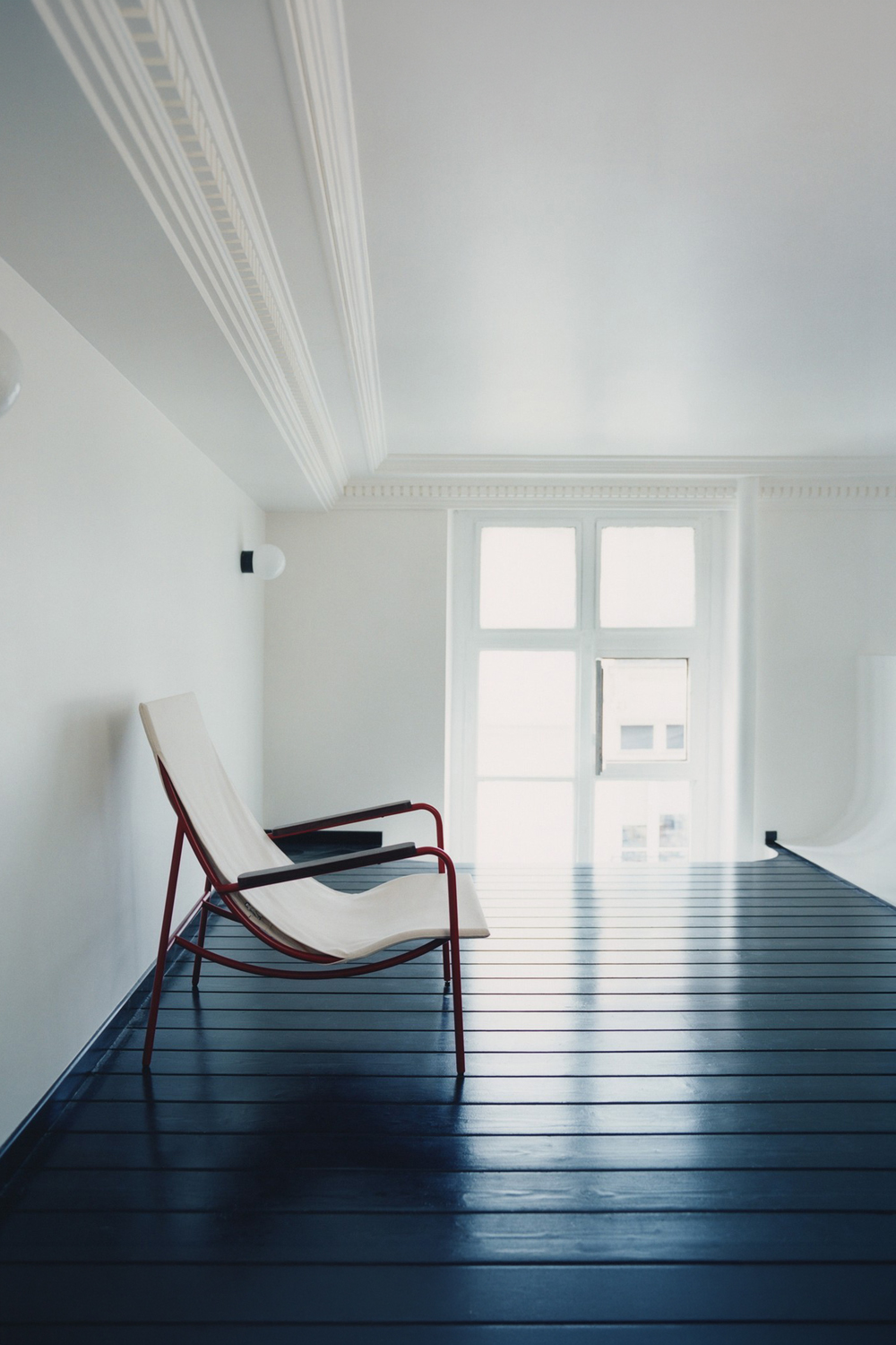 Tropico chair with a red tube frame and cream canvas sling, on a dark blue painted floor beside a tall white window.