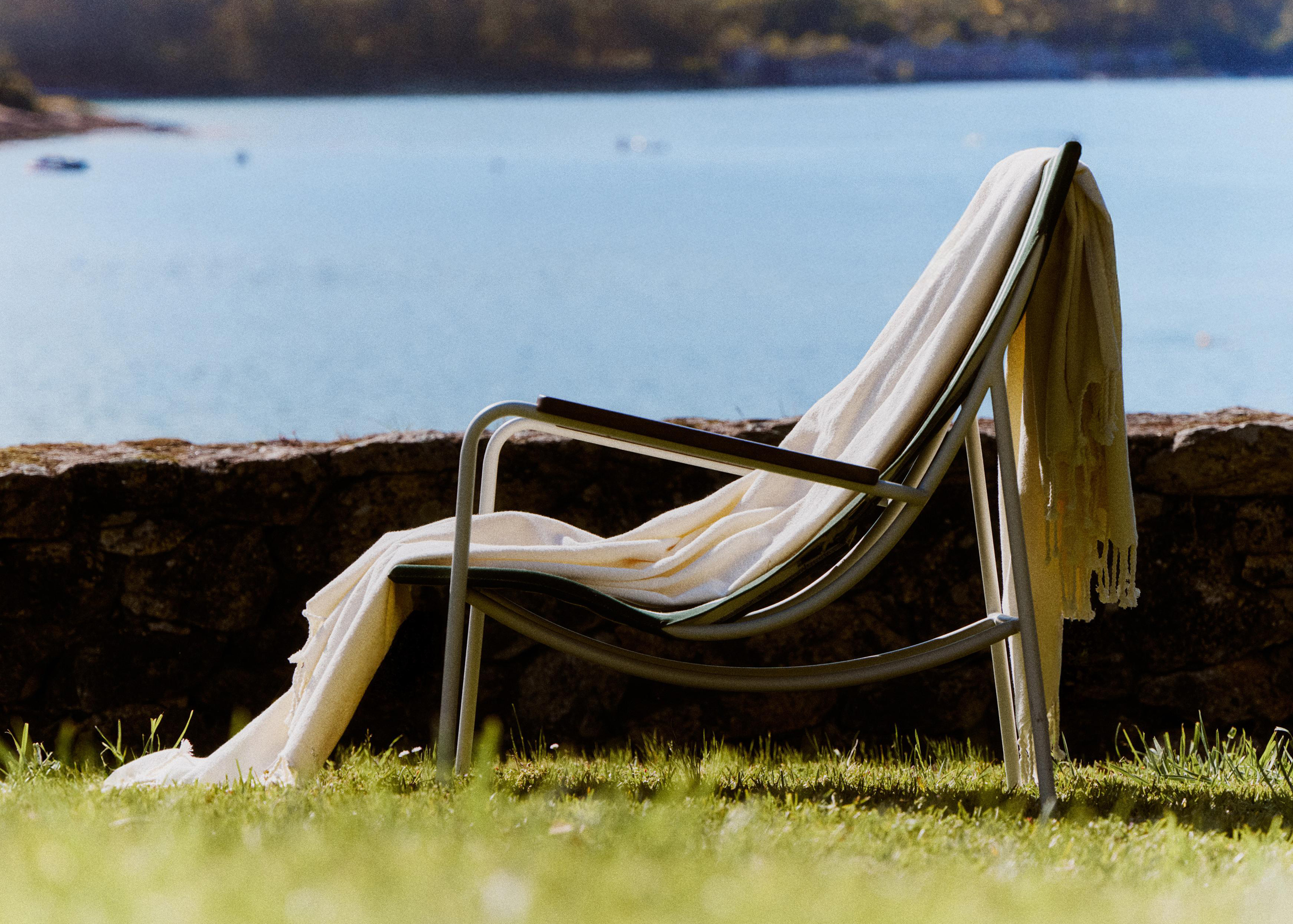 Side view of a white-framed Tropico chair with a cream blanket draped over its back, in front of a stone wall and the sea.