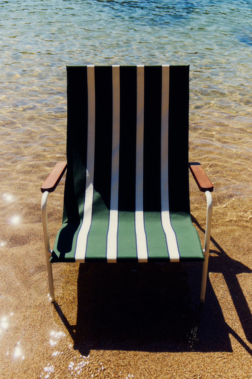 Front view of a Tropico chair with green and cream striped canvas and a white frame, standing in shallow water on a sandy beach.