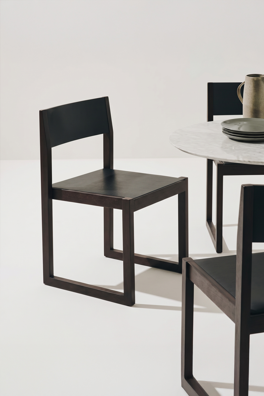 Ray chair with a dark wood sled-base frame and a black leather seat and backrest, beside a round white marble table set with plates and a jug.