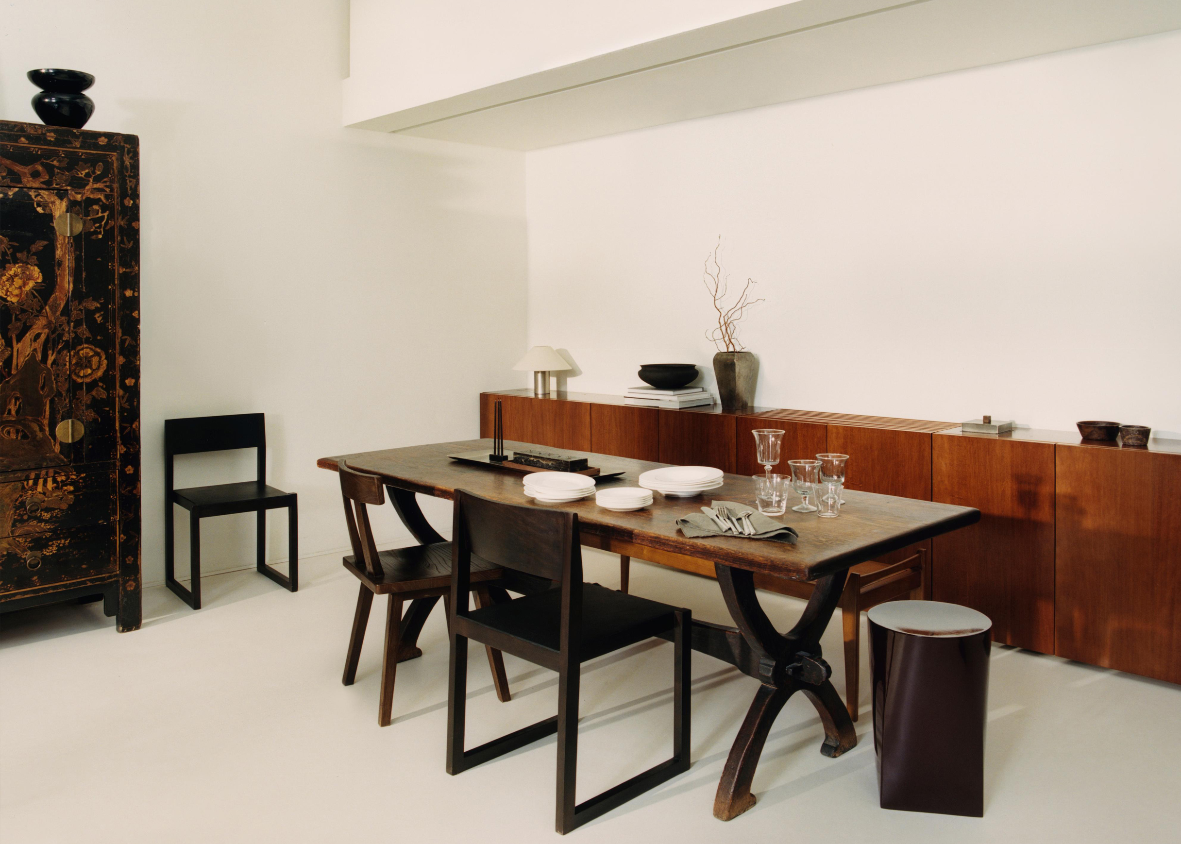Ray chair at a worn wooden trestle table set with white plates and glasses, with a wooden sideboard and a lacquered cabinet behind.