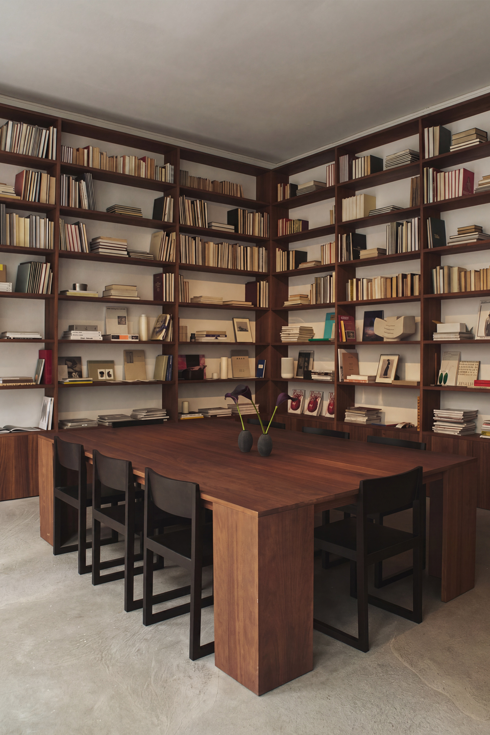 Ray chairs around a long reddish-brown wooden table, in front of a corner of built-in wooden bookshelves.