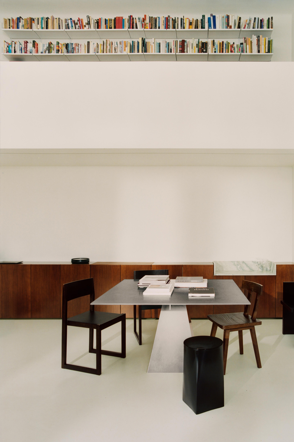 Ray chairs at a grey metal table stacked with books, beside a wooden chair and a black stool, in a white room under a high bookshelf.