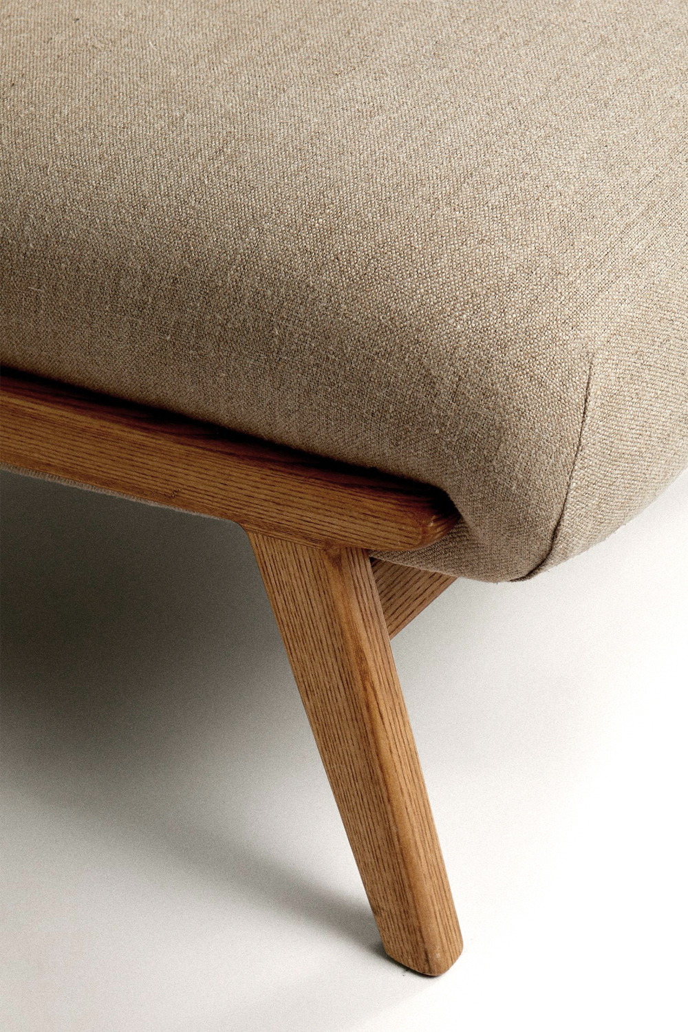 Close-up of the beige linen seat edge, with its fine weave over a light oak frame and a tapered leg.