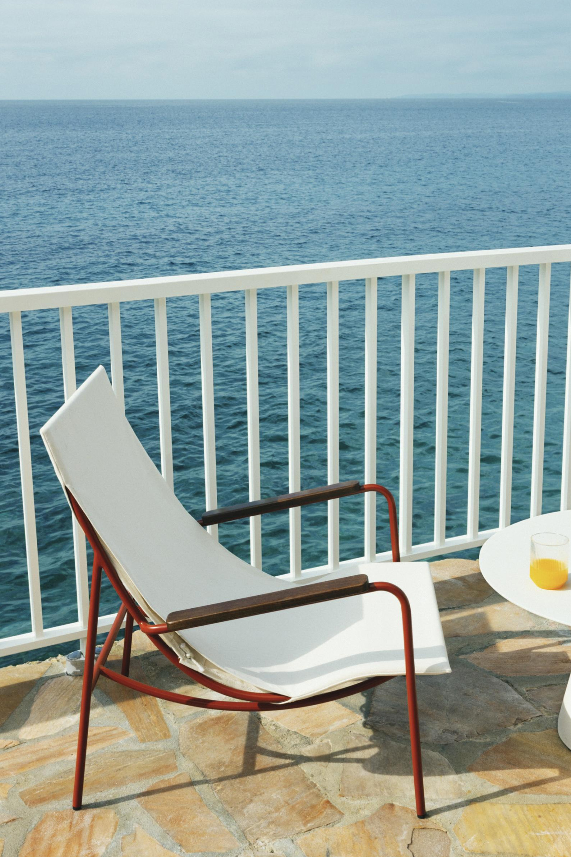 Red-framed Tropico chair on a stone terrace, next to a white railing and a small table with orange juice, overlooking the sea.