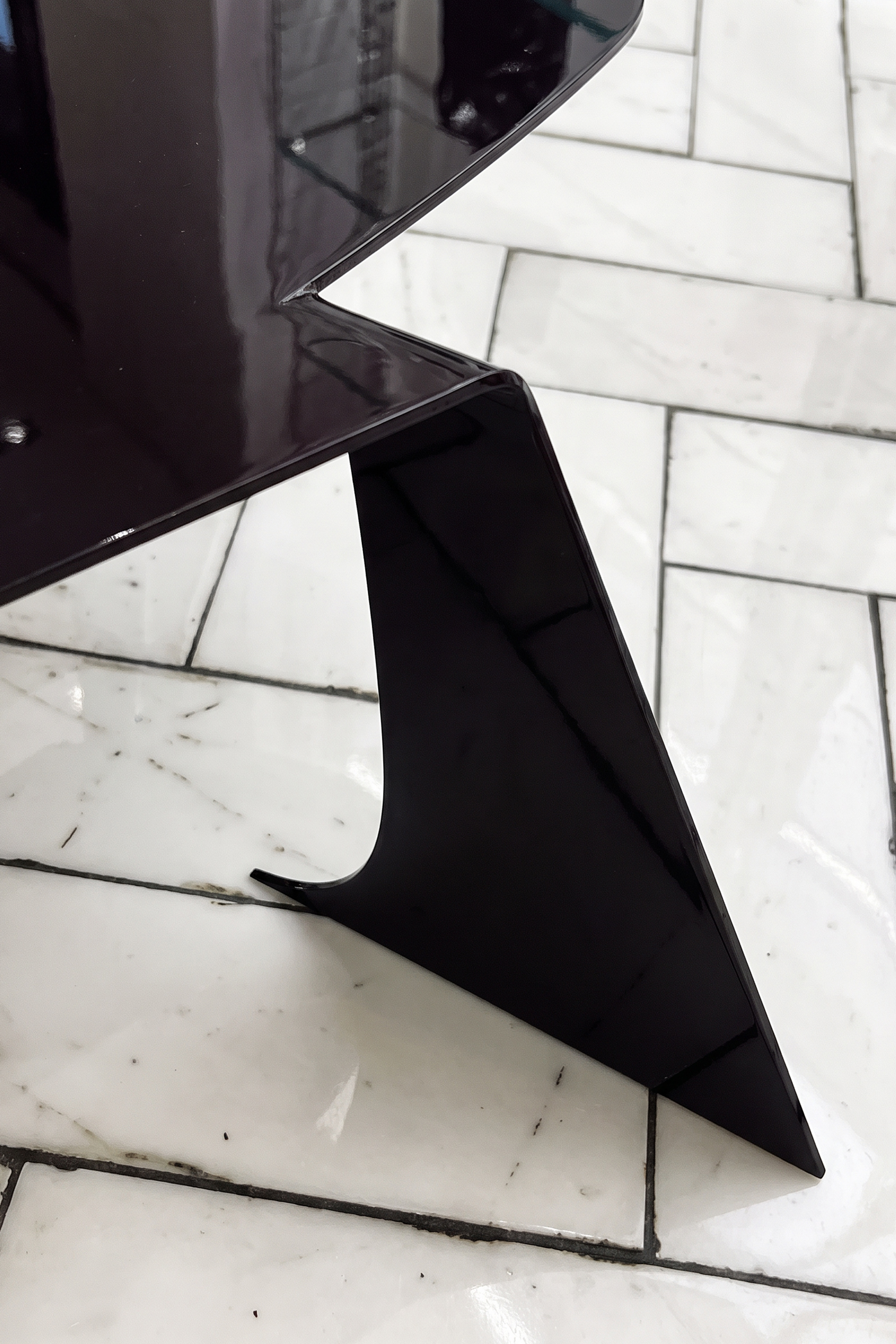 Close-up of the Bend table's glossy dark aubergine steel leg, a folded triangular plane with a curved cut-out, on a white herringbone marble floor.