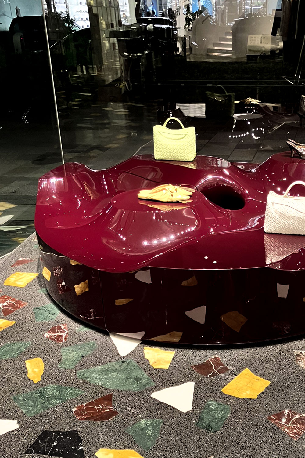 Sun Disc table in a dark Bottega Veneta window, with a lime-green handbag, a yellow shoe and a white bag on its glossy burgundy surface, above a terrazzo floor.