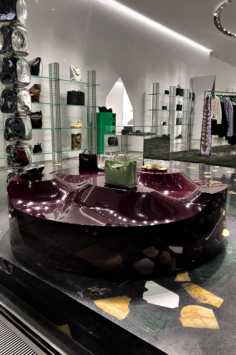 Sun Disc table in a Bottega Veneta Store, holding a green handbag and shoes, with glass shelves and clothes rails in the background.
