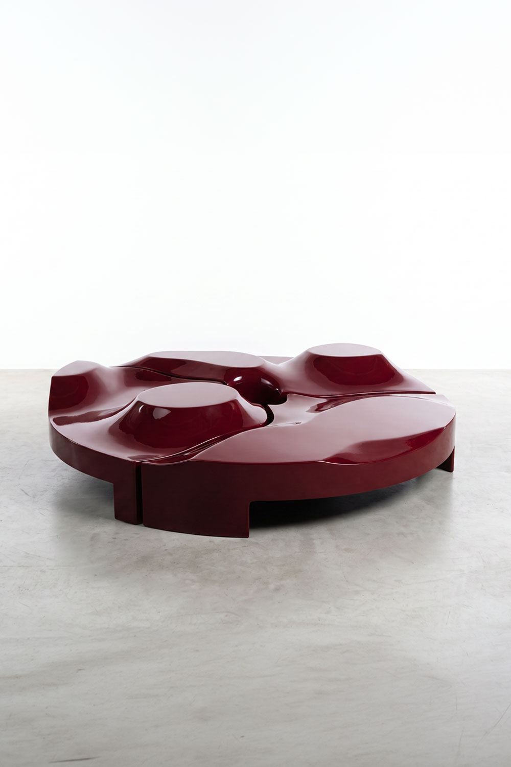 Glossy burgundy Sun Disc table seen from a low angle, with a wavy top, three raised round platforms and short block legs on a concrete floor.