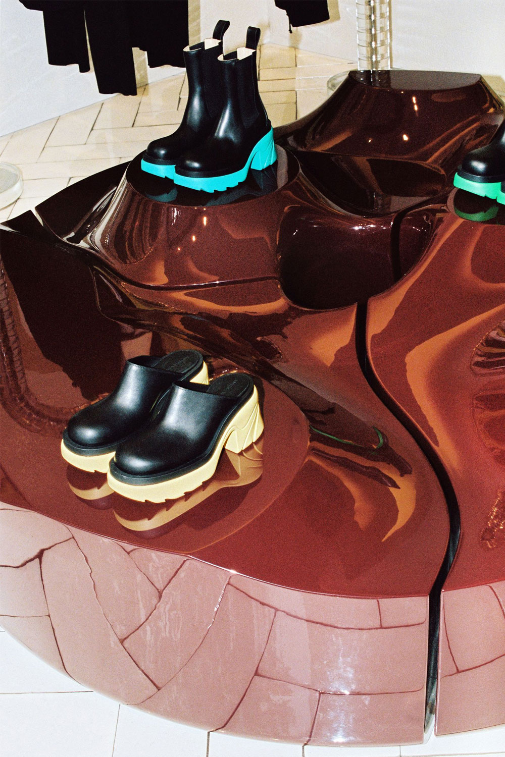 Glossy burgundy Sun Disc display table with raised round platforms, showing teal-soled black boots and black mules with pale yellow soles.