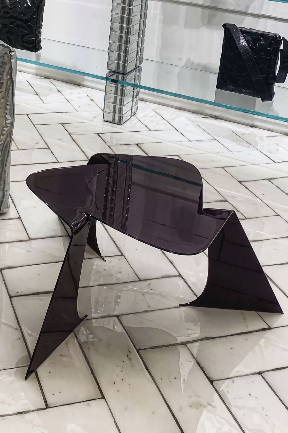 Bend table in glossy dark aubergine, seen from above and to the side, with a rounded triangular top and three folded, angular legs on a marble herringbone floor.