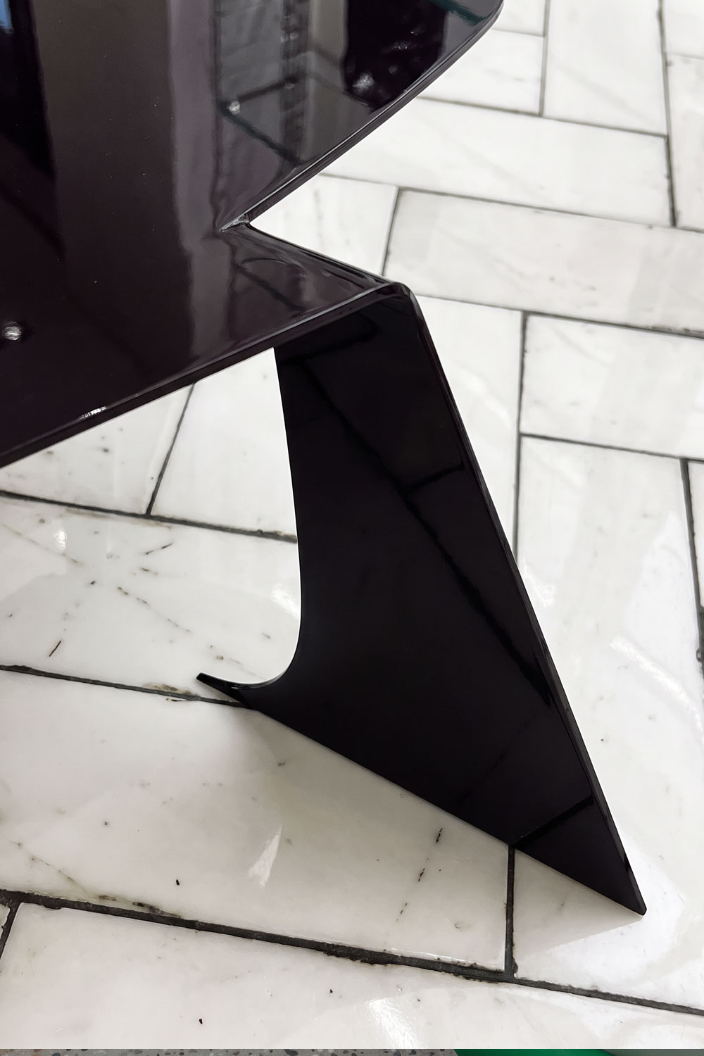Detail of one Bend table leg, where the glossy top folds down into a sharp triangular plane with a curved notch, on white marble tiles.