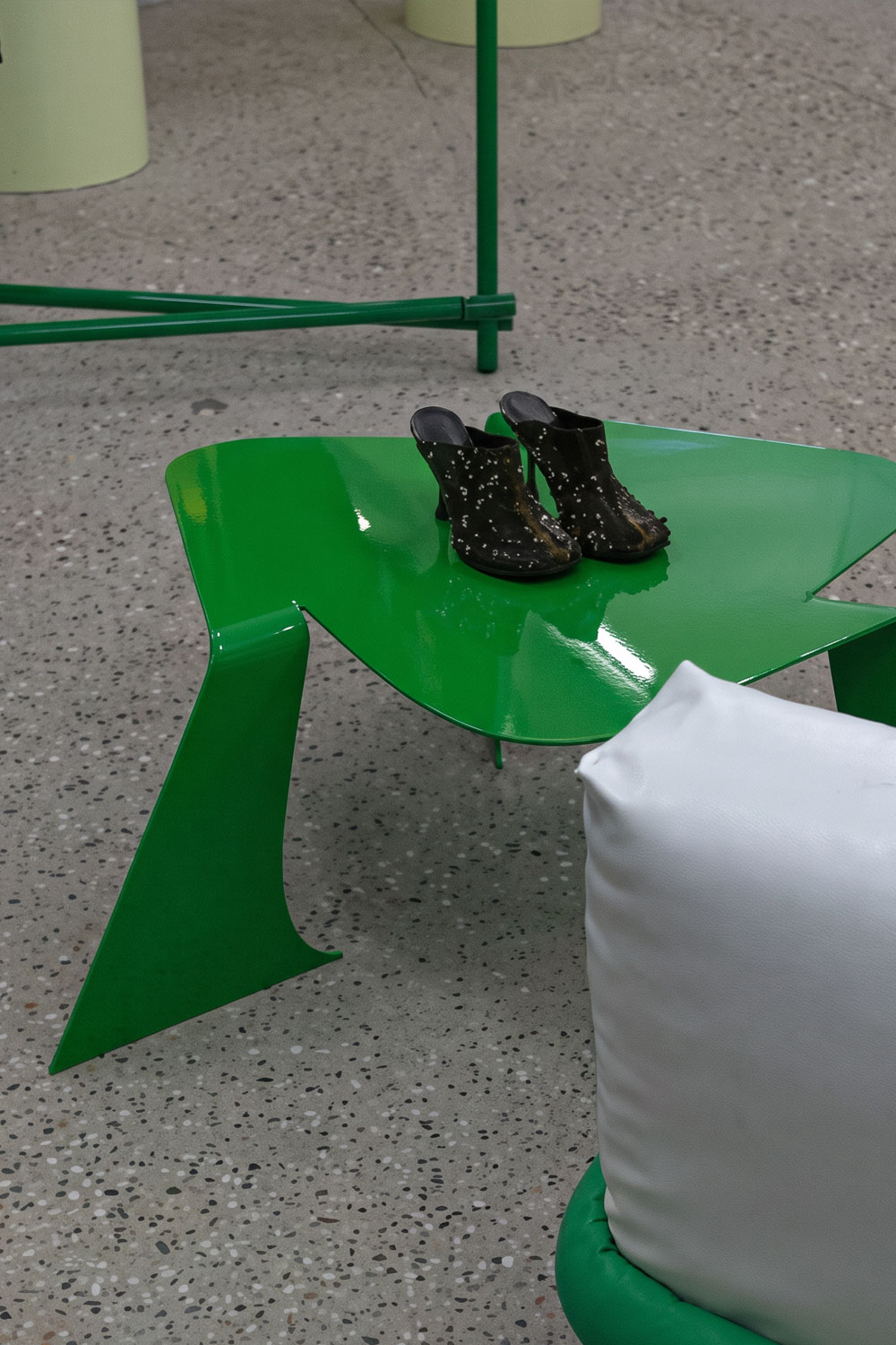 Bright green Bend table in a store, with a pair of black studded heeled mules on its glossy top, beside a white padded seat and a green rail.