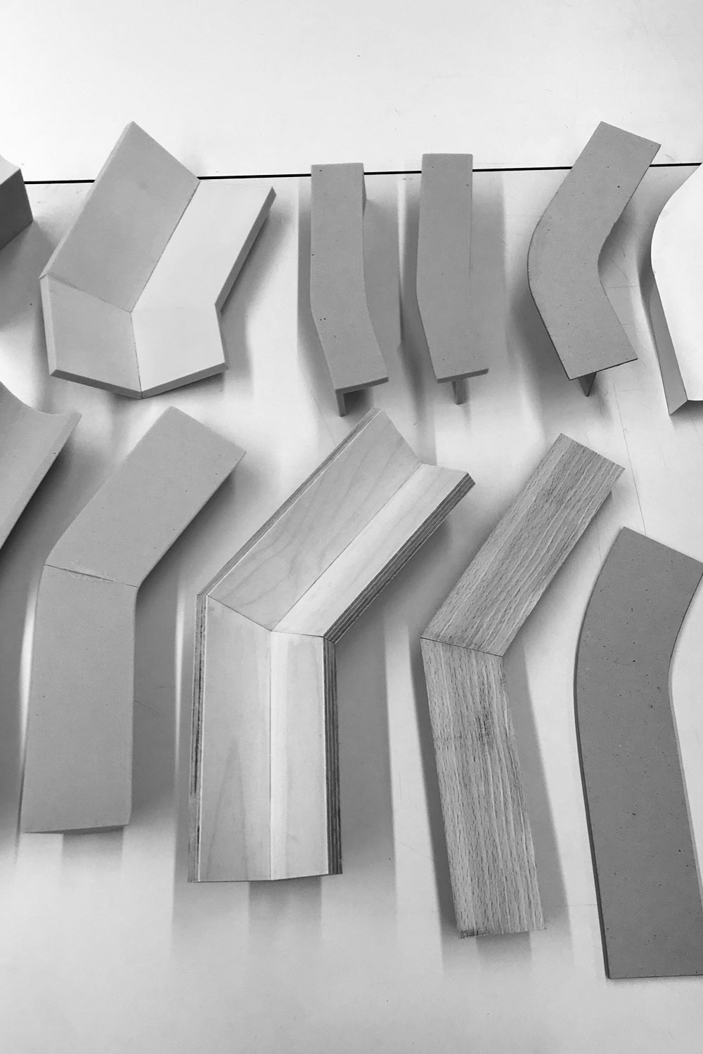 Black and white photo of grey and wooden models of bent tray shapes, laid out on a white surface