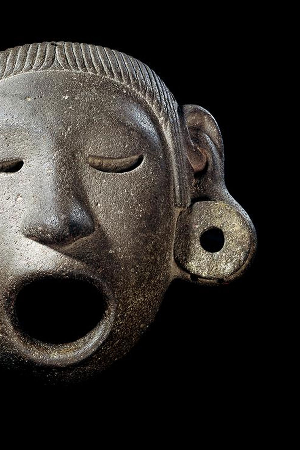 Pre-Hispanic stone mask of Xipe Tótec, with closed eyes, open mouth and a round ear spool