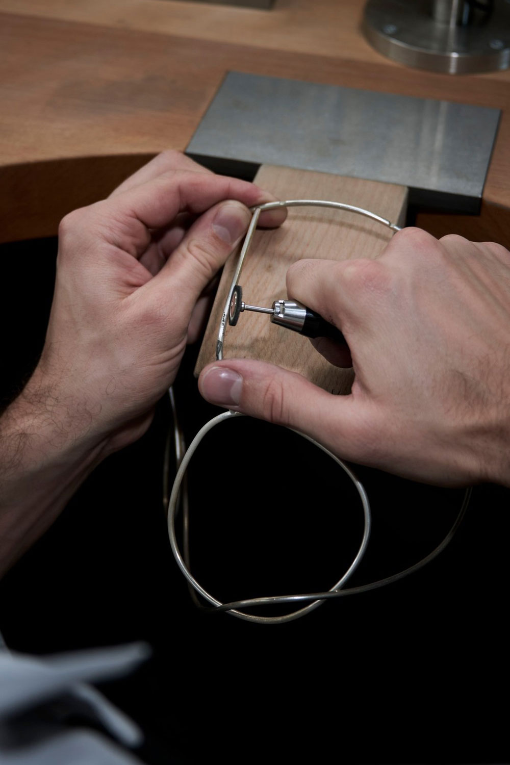 Jeweller's hands shaping a thin silver wire frame against a wooden block with a small tool, at a workbench.