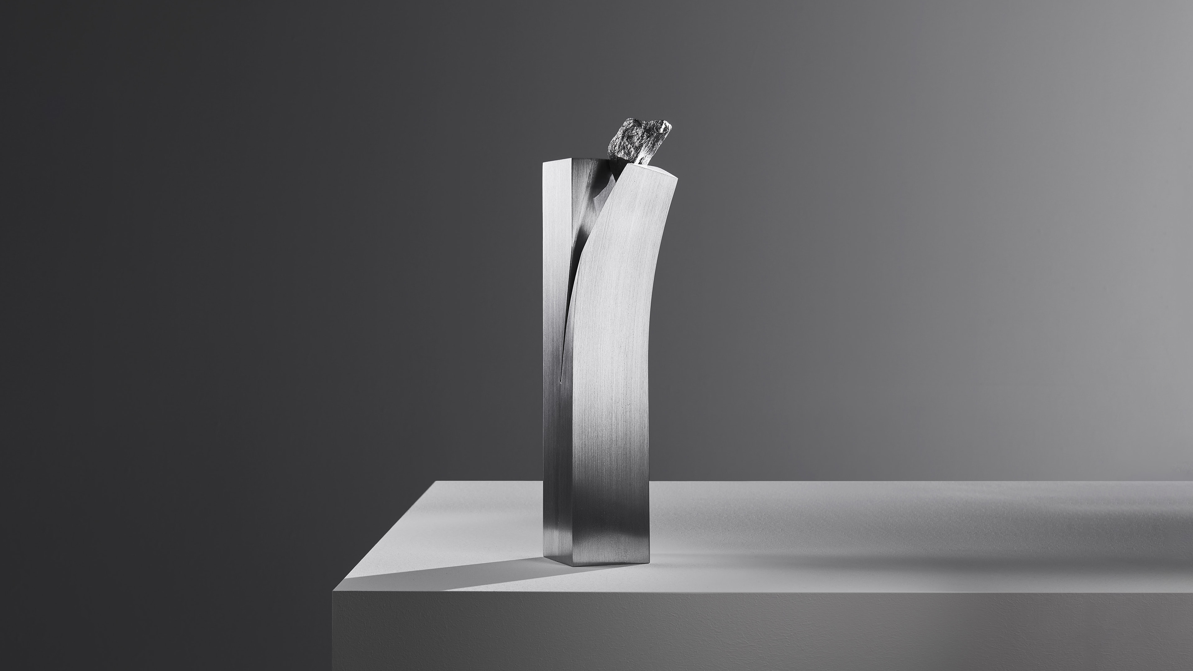 Two tall brushed metal slabs, one straight and one curved, with a dark rough rock wedged in the gap at the top, against a grey gradient.