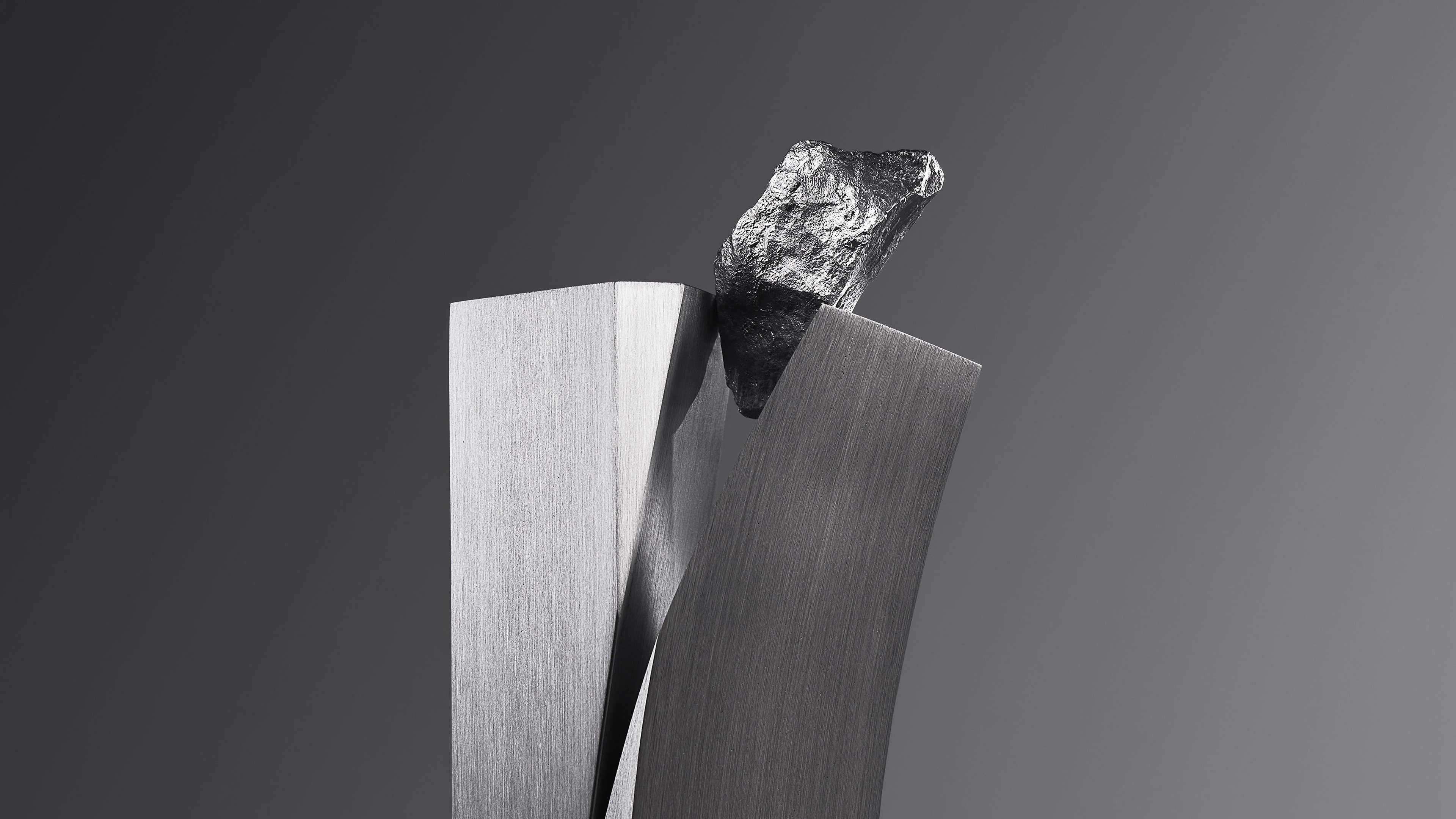 Full view of the sculpture: a slim brushed metal column split by a curved slab, a rough rock on top, standing at the corner of a pale table