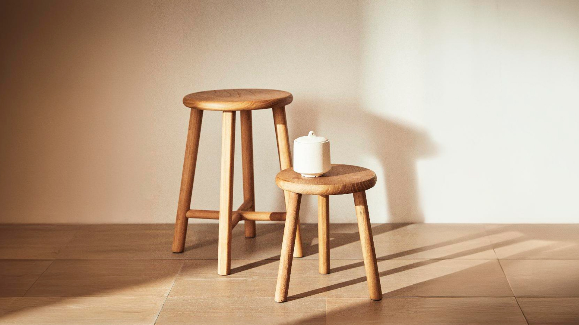 Two Teide stools side by side on a tiled floor in strong sunlight, a tall one with a cross stretcher and a small one holding a white lidded ceramic jar.